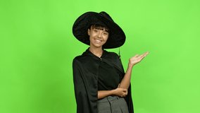 Young woman wearing witch hat extending hands to the side and inviting to come over isolated background. Green screen chroma key