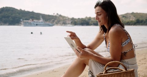 Caucasian happy female relaxing on beach by the ocean reading book on digital tablet 