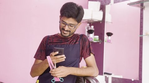 Happy smiling Indian barber busy using mobile phone at saloon - concept of internet, social media and technology