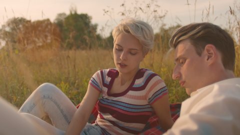 Beautiful young couple chat and joke, lay on picnic blanket in picturesque summer field. Millennial boyfriend and girlfriend on romantic date. Cinematic summertime afternoon. Authentic smiles 
