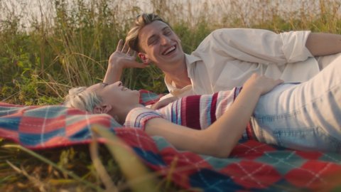 Beautiful young couple relax and chat on picnic blanket in summer field. Romantic date for two lovers on lazy hot spring day. Millennials spend time in nature. Flirting and smiles, sign of affection