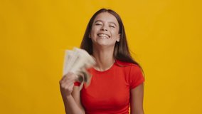 I'm Rich. Joyful Woman Holding Money And Throwing Dollar Banknotes Posing Under Falling Paper Currency Smiling To Camera Over Yellow Studio Background. Financial Profit, Big Luck Concept