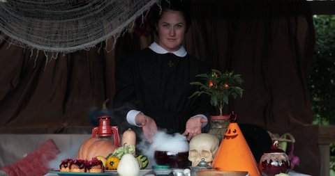 Woman in Halloween nun costume shows smoky potion in glass bowl with Halloween decorations Jack lantern and skull on table. Mystery gothic lady preparing magic potion for Halloween witchcraft