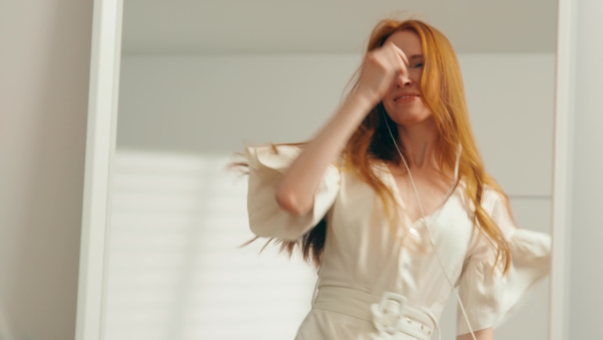 The general plan: A young attractive girl with red hair is dancing merrily at home in front of a mirror with headphones. A beautiful woman in a white dress admires the reflection, dancing. Slow mo, 4K Royalty-Free Stock Footage #1080390143
