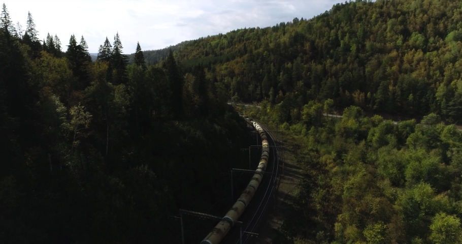 Freight long train carries with oil tank and petrol carriages an electric locomotive by Trans Siberian railways under the rock and near mountain river. Aerial drone wide view at summer sunny day Royalty-Free Stock Footage #1080391535