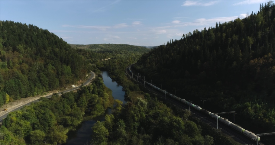 Freight long train carries with oil tank and petrol carriages an electric locomotive by Trans Siberian railways under the rock and near mountain river. Aerial drone wide view at summer sunny day Royalty-Free Stock Footage #1080391538