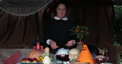 Woman in Halloween nun costume shows smoky potion in glass bowl with Halloween decorations Jack lantern and skull on table. Goth lady preparing magic potion for Halloween witchcraft. Slow motion