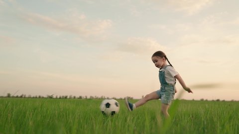 little kid plays football at sunset, baby runs around green football field and kicks ball, child runs to floor in meadow and smiles, learns how to kick ball, dream of becoming team footballer