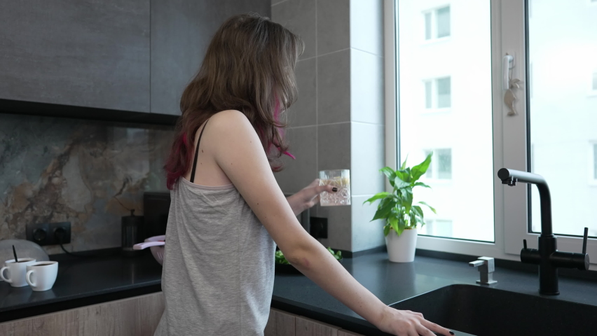 Young woman standing looking out of her apartment kitchen window with a glass of fresh water and lemon in her hand in a close up side view