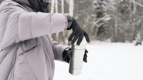In the video, female hands in black gloves pour hot coffee from a thermos into a mug. Pine trees, snow, winter in the background.