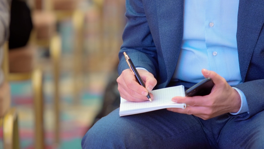 male hand writing in paper notebook with black ballpoint pen, medium close-up shot. a man in a blue business suit makes notes on his knee holding a smartphone in his hand