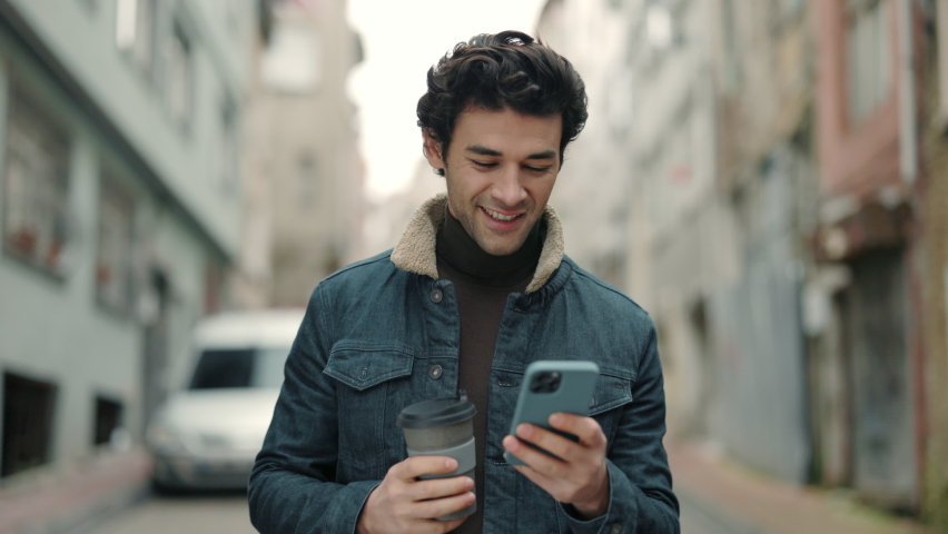 Cheerful young guy walking on city street with cup of coffee and modern smartphone in hands. Handsome arabian man reading some good and smiling. Technology for lifestyles. | Shutterstock HD Video #1080415184