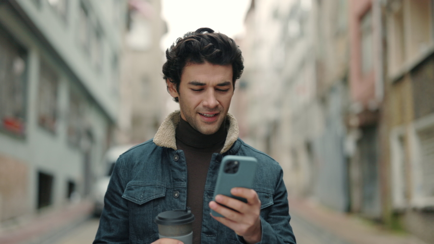 Cheerful young guy walking on city street with cup of coffee and modern smartphone in hands. Handsome arabian man reading some good and smiling. Technology for lifestyles. Royalty-Free Stock Footage #1080415184
