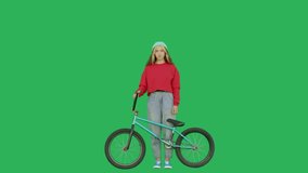 Sporty girl wearing sweatshirt and knitted hat standing with bmx bike over green screen background. Pretty young woman holding bicycle looking at camera on chroma key. 4k raw video footage