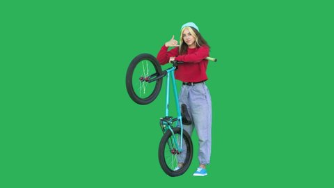 Sporty girl wearing sweatshirt and knitted hat holding bmx bike over green background. Pretty young woman with bicycle in her hands looking at camera showing shaka sign. Chroma key