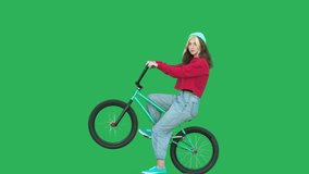 Sporty girl wearing sweatshirt and knitted hat sitting on bmx bike over green background. Pretty young woman on bicycle looking at camera. Chroma key. 4k raw video footage