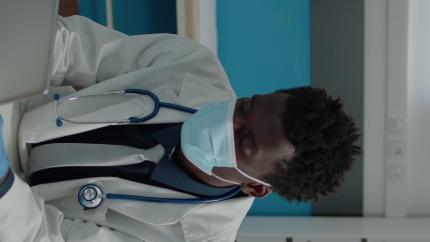 Vertical video: Close up of young medic with face mask in medical cabinet using modern laptop on desk. Doctor sitting with digital device typing on keyboard while wearing stethoscope for healthcare, a Technology Video by DC Studio