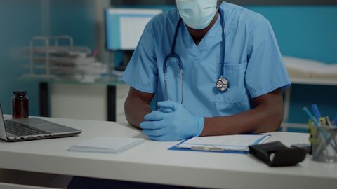Portrait of man with nurse occupation looking at camera with face mask and uniform. Medical assistant sitting at desk in healthcare office with laptop and professional instruments