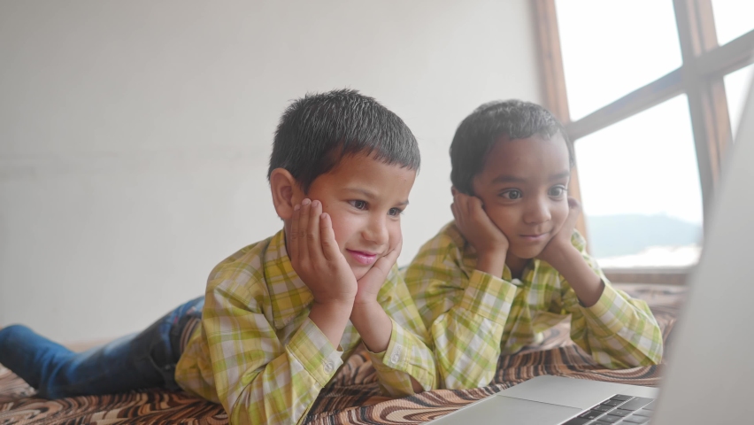 Two Indian Asian school kids wearing school uniforms are engrossed and attending online video classes using a laptop in an indoor house setup. Remote learning and education concept.  Royalty-Free Stock Footage #1080422330
