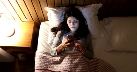 Woman lying in bed at night looking at phone. Person staring at blue cellphone screen before sleep