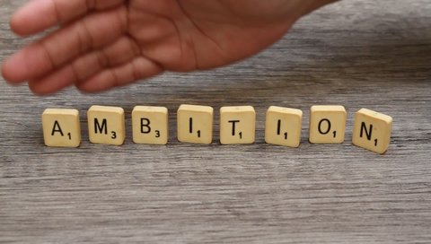 Jakarta, Indonesia - September 16th, 2021: A hand sweeping the word "AMBITION" from the Scrabble Game Letters on the wooden table