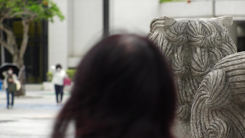 NAHA, OKINAWA, JAPAN - AUG 2021 : People  wearing surgical masks to protect from Coronavirus (COVID-19). View around central downtown area, Kokusaidori street. Camera focus on Shisa statue.