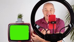 middle-aged charming man, 60-65 years old blogger sits in front of a ring light and a red smartphone, emotionally talks on the topic of business, politics, old TV with a green screen for video