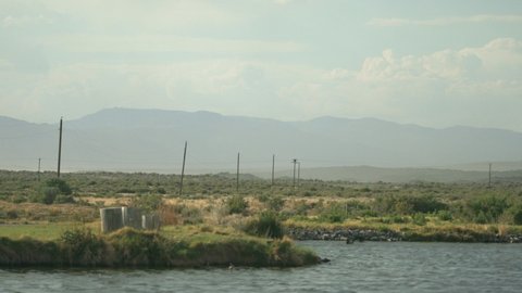 Peaceful telephoto footage of a pond and telephone poles going into the distance, large clouds and mountains are seen in the distance, with sagebrush and other dry grasses.