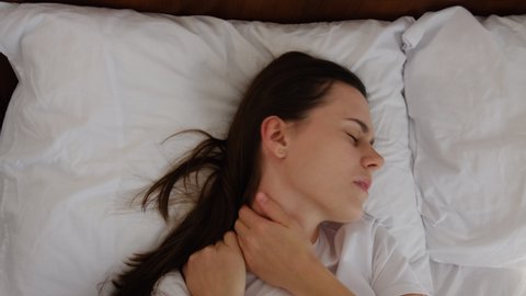 Top view of tired young brunette woman lying on bed stretching and rubbing stiff shoulder muscles after waking up in morning. Upset girl feels discomfort because of bad mattress or posture problems
