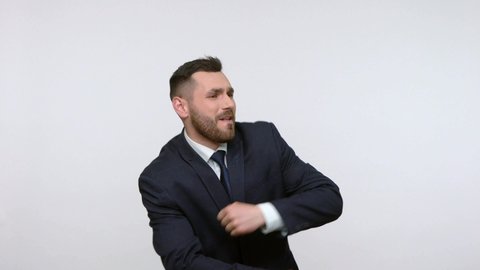 Extremely happy bearded businessman in black official style suit dancing and moving to rhythm, expressing positive emotions, celebrating. Indoor studio shot isolated on gray background.