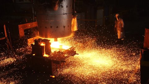 Laborer in protective suit stands by tank with liquid metal and bright sparks spread in contemporary steelmaking plant workshop