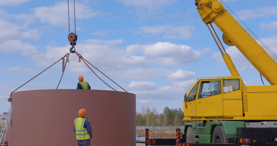 A team of workers in overalls and helmets help a large yellow truck crane move huge metal parts on a nearby truck. | Shutterstock HD Video #1080463337