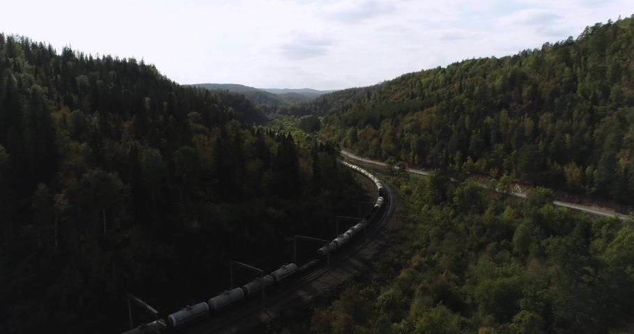 Freight long train carries with oil tank and petrol carriages an electric locomotive by Trans Siberian railways under the rock and near mountain river. Aerial drone wide view at summer sunny day Royalty-Free Stock Footage #1080468440