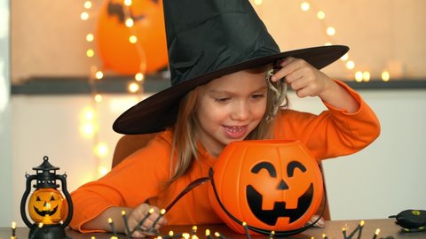 Halloween. Happy little girl in witch costume and black hat fun brews potion from spiders, snakes, frogs in pumpkin pot.