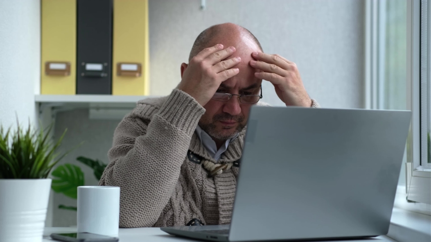 Middle-aged serious male is looking at laptop, taking off glasses, tired while sitting at table. Working at home office. Closes laptop, stops work. Remote job, freelance, business.