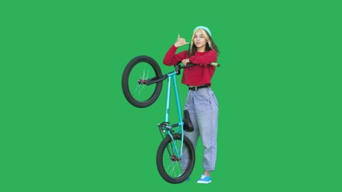 Sporty girl wearing sweatshirt and knitted hat holding bmx bike over green background. Pretty young woman with bicycle in her hands looking at camera showing shaka sign. Chroma key