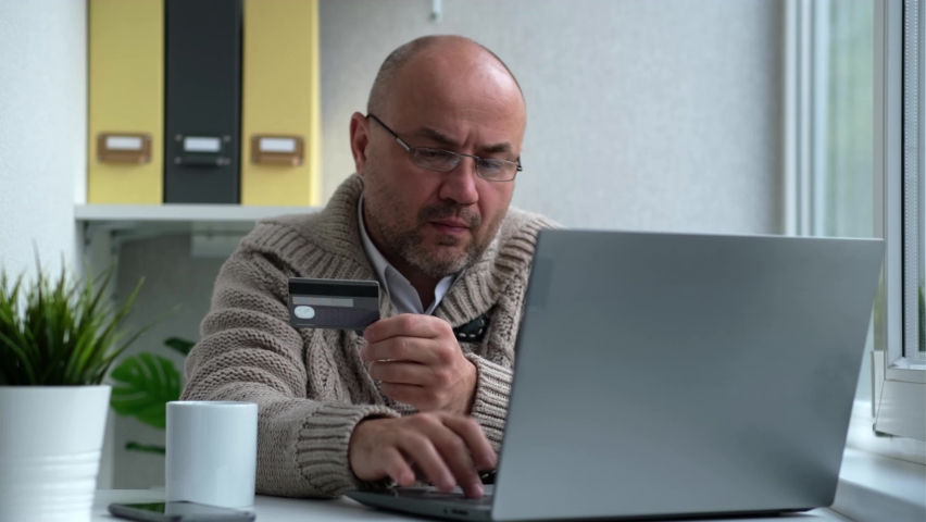 Serious man wearing glasses shopping online, paying with credit card while making orders via the Internet. Successful businessman making transaction using mobile bank application or online store.