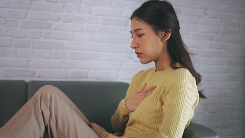 Asian young woman feeling discomfort as suffering from heartburn holding chest with closed eyes and sitting with folded legs on couch at home Royalty-Free Stock Footage #1080473603