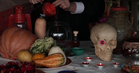 Hands preparing magic potion and pouring hot dark liquid from jug. Witch make potion in glass bowl with Halloween decorations, pumpkins, candles and skull on table. Halloween magic potion making