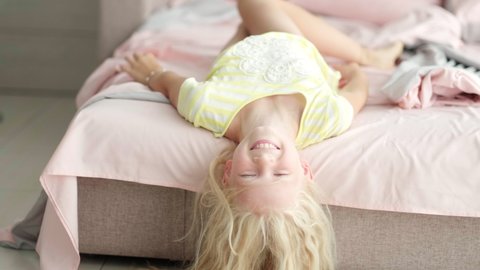 Portrait handsome cute little blonde girl having fun in bed in the morning. Adorable kid spinning, laughing, lying upside down.