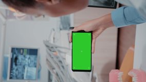 Vertical video: Woman holding smartphone with horizontal green screen at oral care clinic. Dentist looking at chroma key with isolated background and mockup template for dental checkup