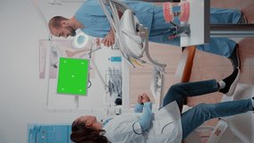 Vertical video: Dentist and assistant working with green screen on display while discussing oral care examination. Woman and man using chroma key with mockup template and isolated background on