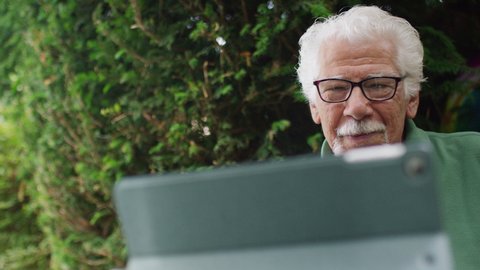 Retired senior male talking on a video call on a digital tablet in his back garden, in slow motion