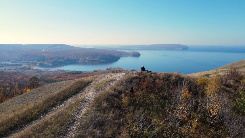 Cinematic aerial view of a beautiful autumn forest with golden foliage, mountain scenery and a lone traveler relaxing sitting on a stone. Dramatic drone shooting in UHD 4K resolution.