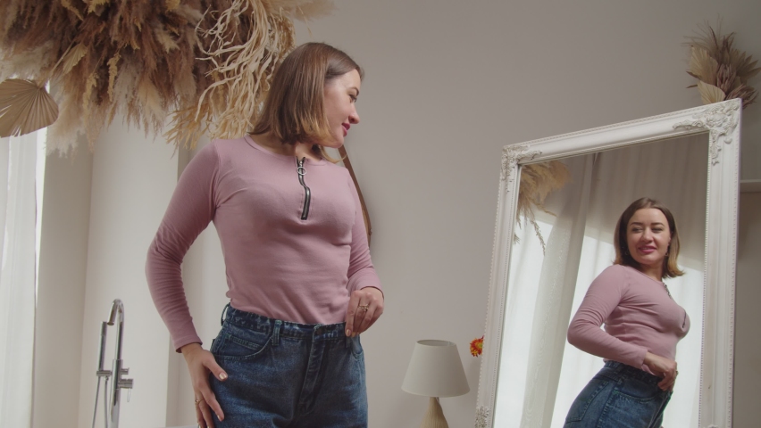 Portrait of cheerful lovely female on oversized jeans checking and celebrating weight loss success with winning gesture, expressing excitement and joy while looking at reflection in mirror indoors. Royalty-Free Stock Footage #1080502097