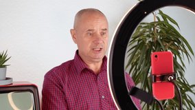 middle-aged charming man, 60-65 years old blogger sits in front of a ring light and a red smartphone, emotionally talks about business, politics and records video, filmed on a 4K camera