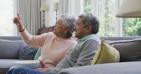Happy biracial senior couple watching tv with popcorn and having fun at home. retirement lifestyle relaxing at home with technology.