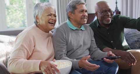 Happy biracial senior couple and african american friend watching tv with popcorn. socialising with friends at home.