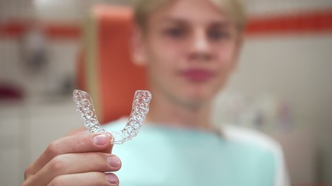 Teenager showing mouthguards and sitting while visiting orthodontist in clinic office spbd. Closeup view of caucasian boy holds transparent sample in hand and showes, looks with smile and sits in