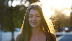 Beautiful woman laughing at camera and touching her long hair in rays of sunset, blurred street on background. Portrait of happy female model outside. Concept of emotion